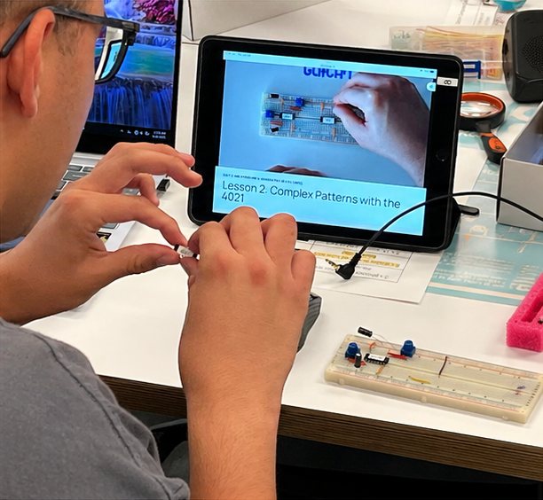 A builder places a part on his breadboard while the lesson video plays on a tablet