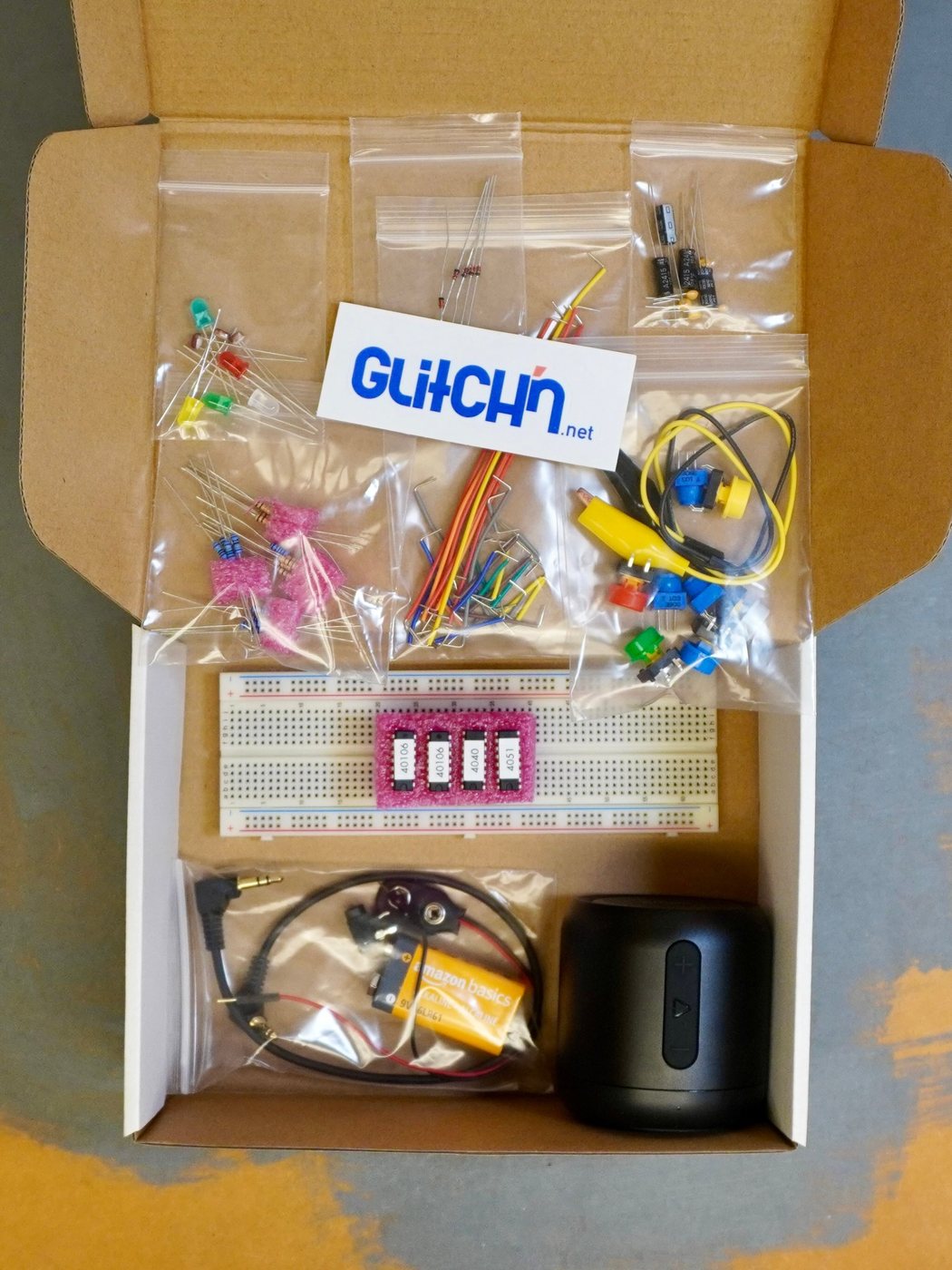An open Glitch'n kit box: a breadboard with four labeled chips, small parts in clear bags, wires, a 9-volt battery and a small speaker